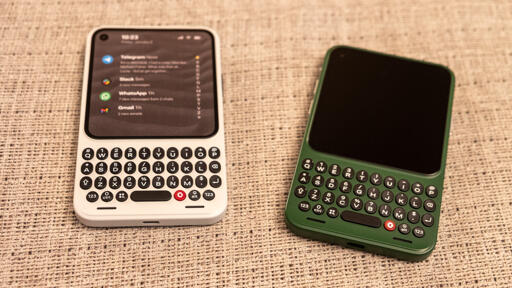 Two Clicks Communicator devices on a table, face up. The left one is white with the screen on showing some notifications and the right one is green with the screen off.